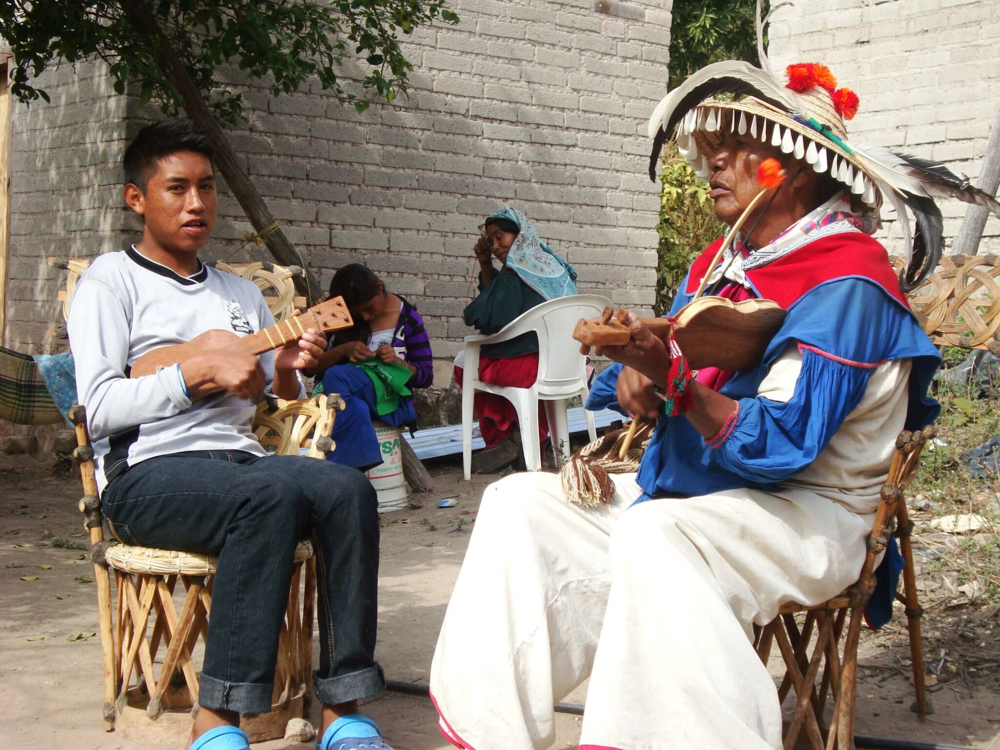 A teenage Huichol boy sits on a chair playing a small stringer instruments. An older Huichol man sits on a chair playing a small stringer instrument.