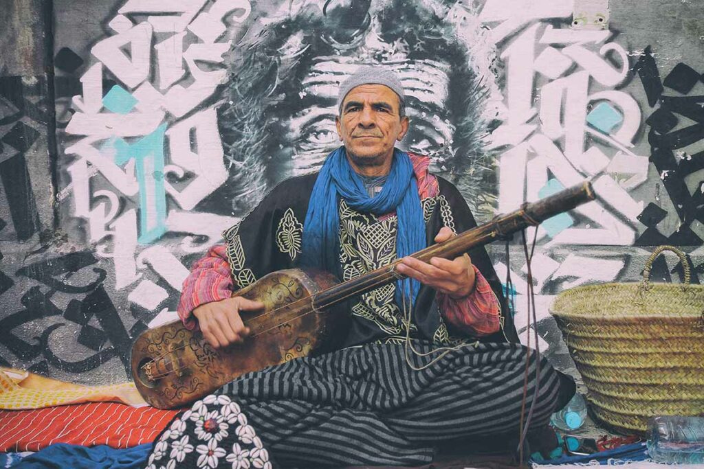 Portrait of a north African man sitting in front of a graffiti wall, playing a traditional instrument.